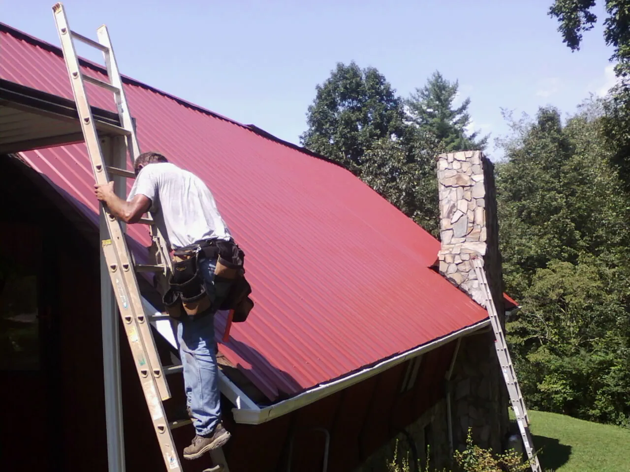 Roofer on a ladder beside a red metal roof and stone chimney — Soffit & Fascia Repair by Roofing Brothers in Hilmar